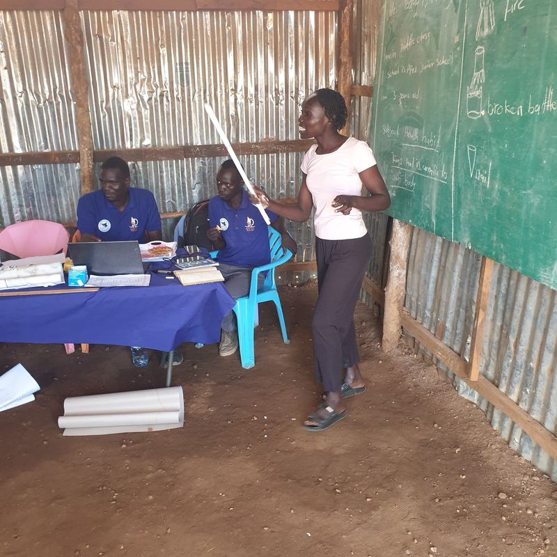 A facilitator leads a teachers' skills workshop in a classroom at Apex Junior Schools, Juba, while two colleagues take notes at a laptop