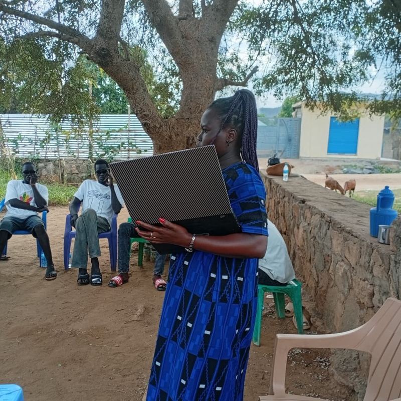 An EFSS volunteer holds up a laptop to residents seated under a tree at a community outreach in Khor Wolliang, Juba